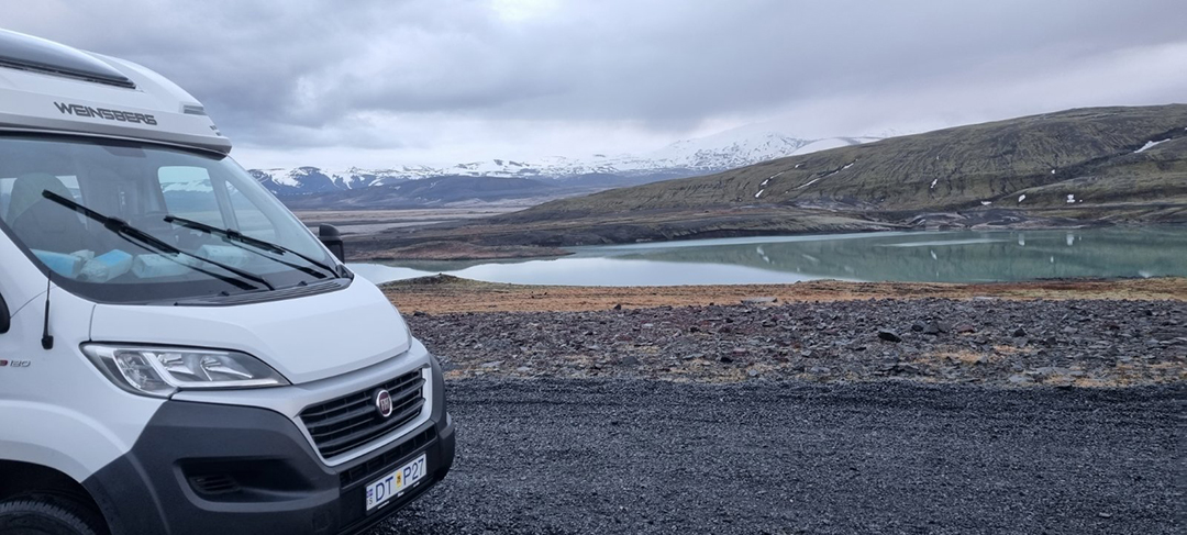 A motorhome parked by a calm Icelandic lake reflecting snowy mountain peaks and peaceful scenery.