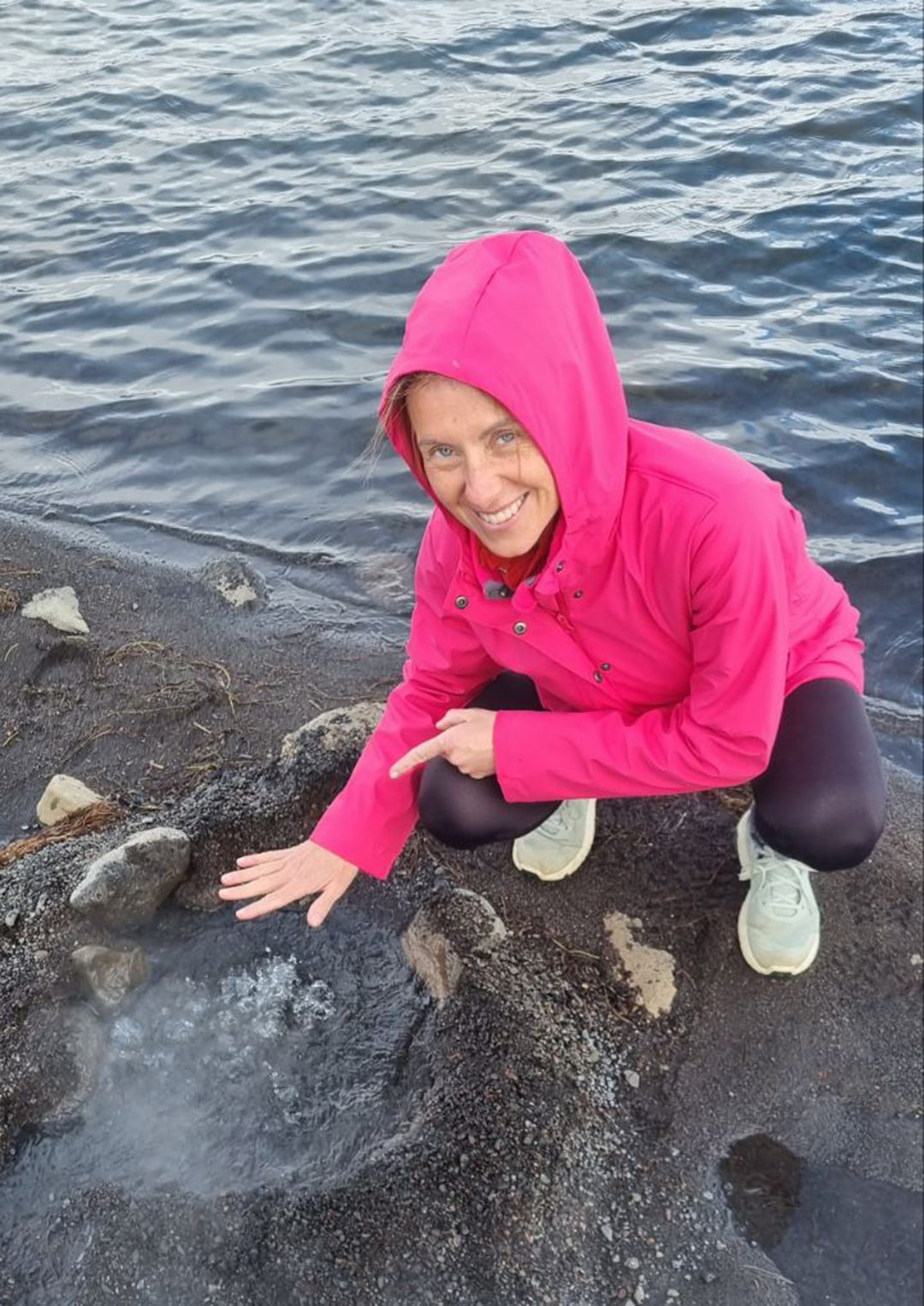 A traveler holding a hand over a small geothermal vent by the lake, with hot steam rising from the ground.