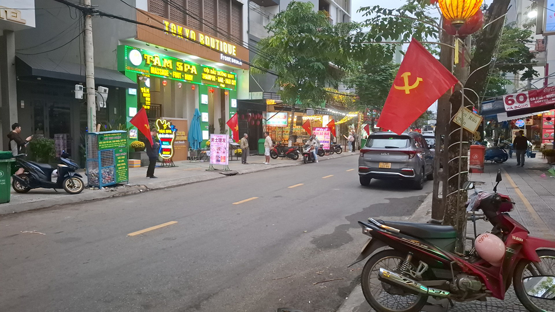 Mestna ulica v Da Nangu v Vietnamu z lokalnimi trgovinami, motorji in vietnamskimi zastavami ob cesti.